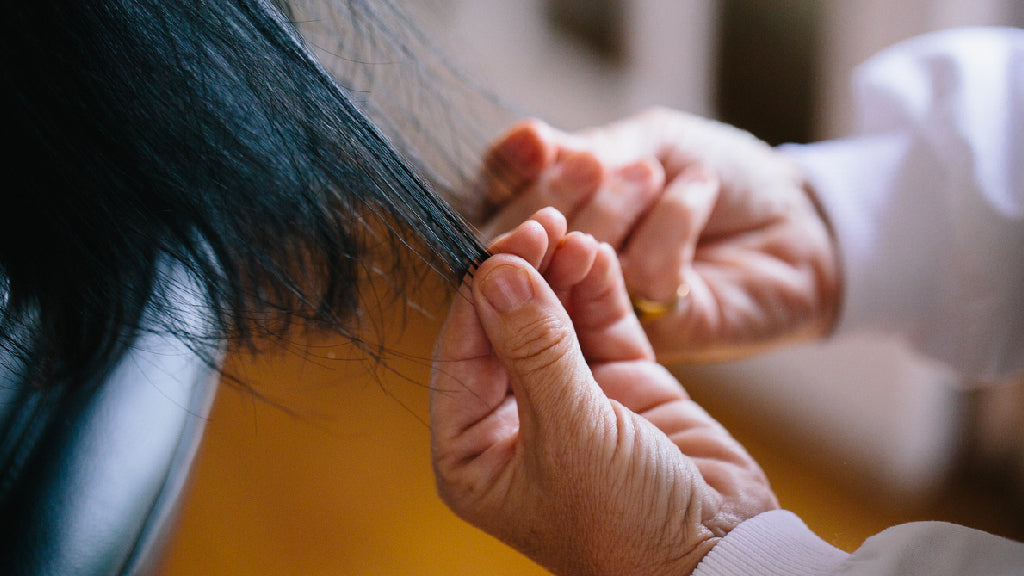 Close up photo showing a white woman's hands examining a woman's long black hair as the woman sits in a glossy black chair