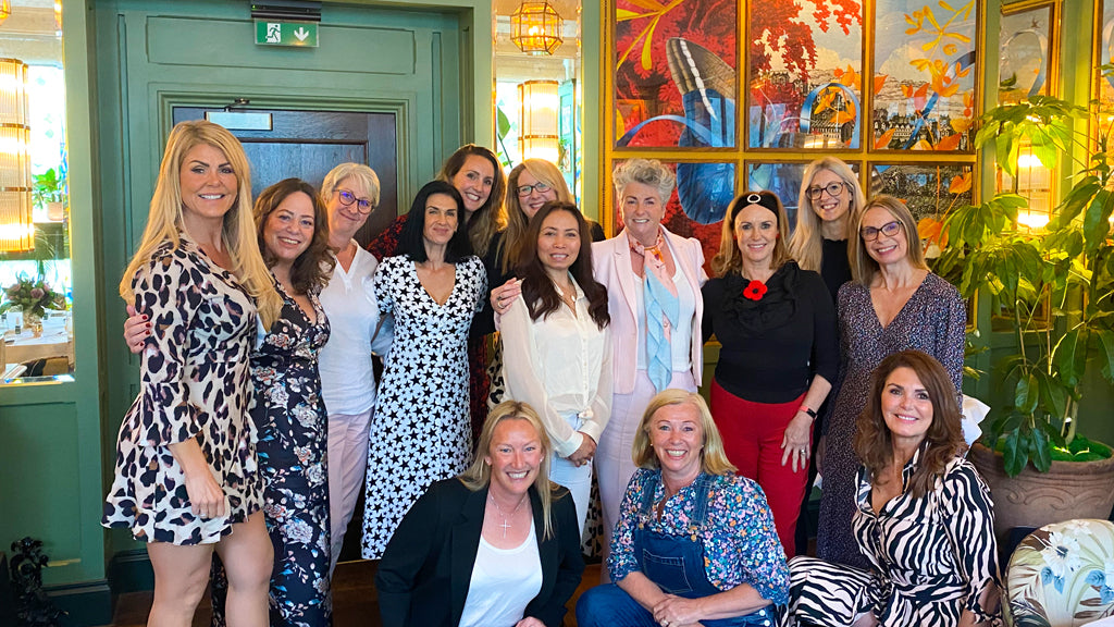 Photo showing a group of women standing inside a brightly coloured room, they are all grouped together wearing pretty dresses and smart clothes and they are all smiling at the camera