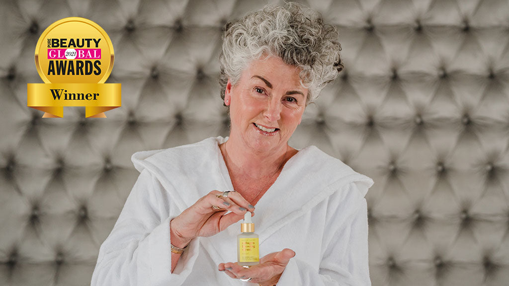 Photo showing Maxine Laceby wearing a dressing gown and holding a bottle of Maxerum, she is sitting against a textured grey headboard and the Pure Global Beauty awards logo has been photoshopped alongside her