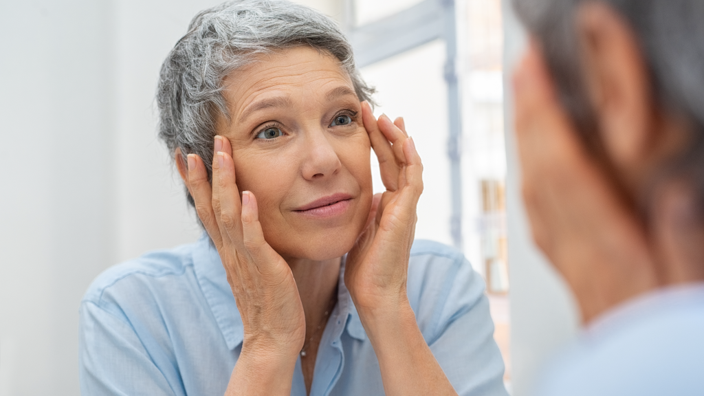 Photo showing a white woman with short grey hair looking at her skin in the mirror