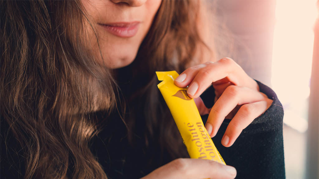 Photo showing a white woman with long brown hair holding an Absolute Collagen sachet to her mouth