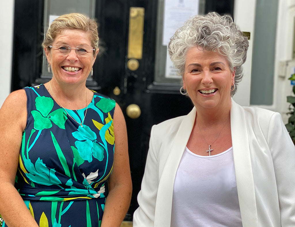 Photo of two smiling white women against a black door, one woman wearing green and blue and one woman wearing white and cream